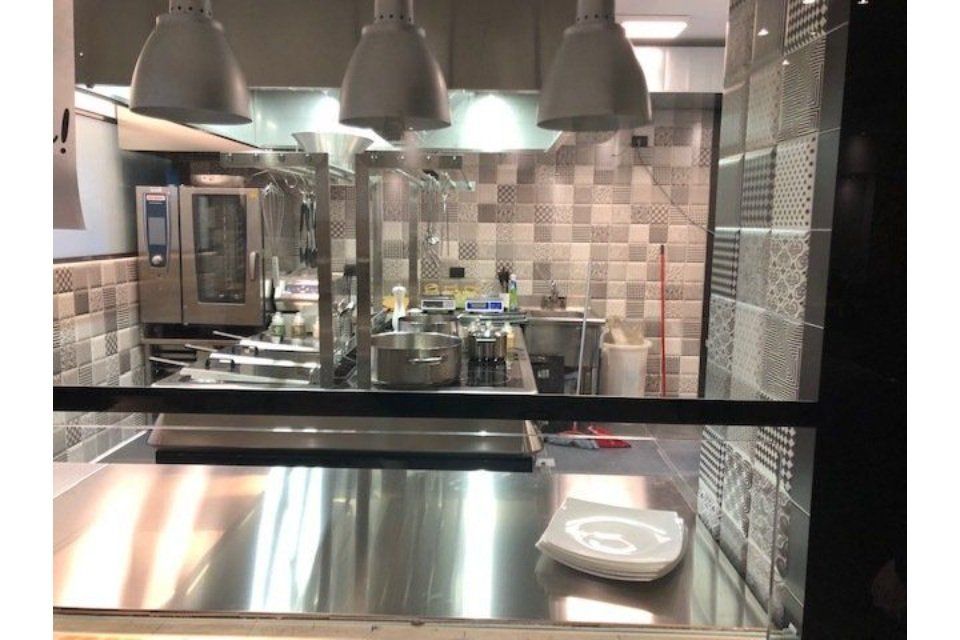 Interno della cucina con banconi e attrezzature in acciaio inox. Luci a soffitto, paraschizzi piastrellato. Piatti vuoti sul bancone.