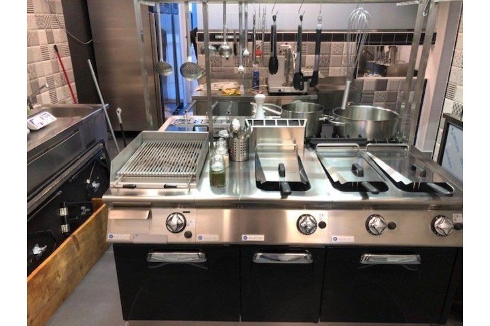 Cucina commerciale in acciaio inox con piastra, friggitrici e utensili appesi sopra.