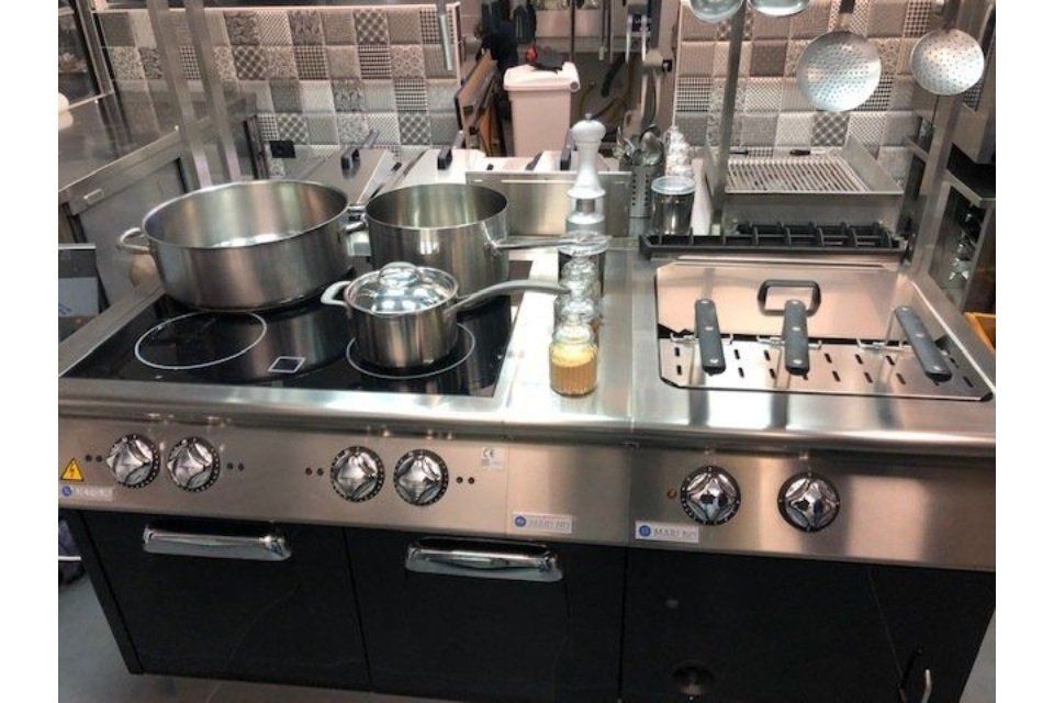 Cucina commerciale con pentole, padelle e friggitrice; acciaio inossidabile, dettagli neri.