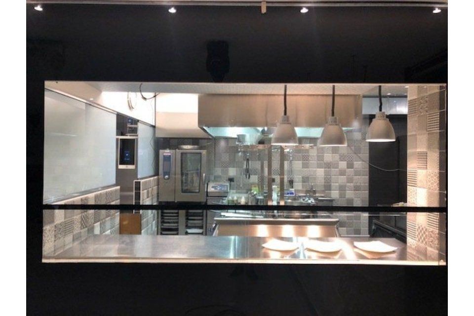 Cucina del ristorante vista attraverso una finestra: banconi in acciaio inossidabile, forno, piano cottura e luci sospese.