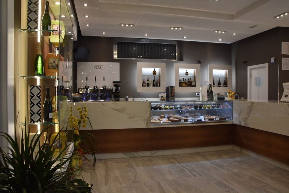 Interno di un bar con bancone in marmo, vetrina, scaffali con bottiglie e piante in vaso.