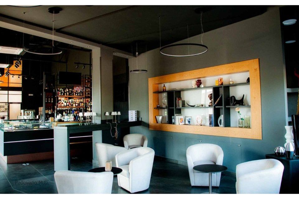 Interno di un bar moderno con area salotto, bar e scaffali espositivi; pareti grigie, sedie bianche, soffitto scuro.