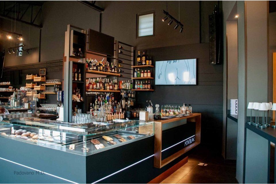 Vista interna di un bar moderno con bottiglie di liquore sugli scaffali, una vetrina e un bancone nero e nei toni del legno.