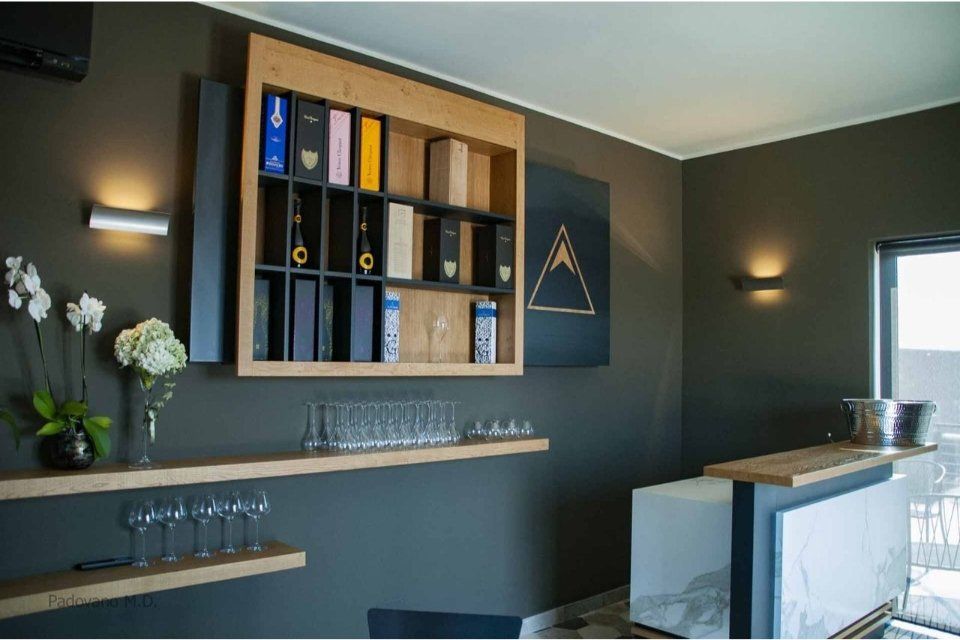 Wine bar con scaffali espositivi, bicchieri e bancone integrato. Pareti scure, dettagli in legno e illuminazione d'atmosfera.