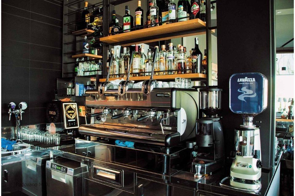 Bar con macchina per caffè espresso, bottiglie di liquore e insegna del caffè Lavazza. Nero e acciaio inossidabile.