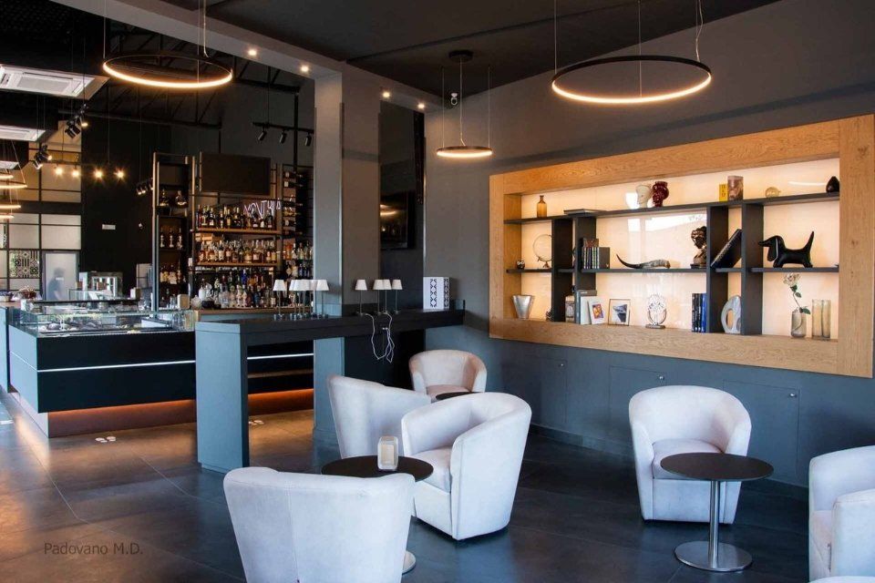 Area lounge con sedie bianche, tavolini e un bar sullo sfondo con scaffali e illuminazione circolare.