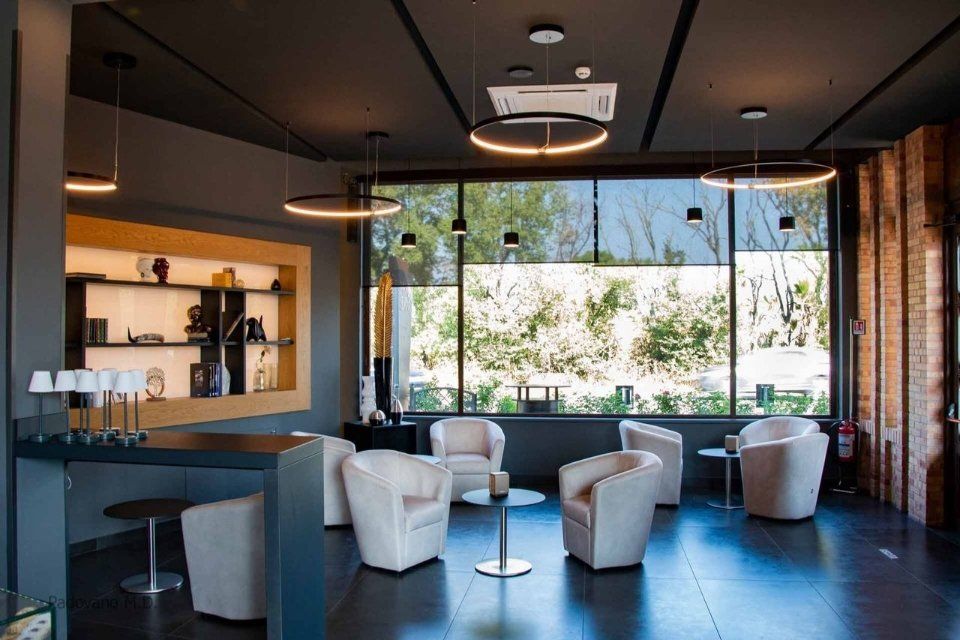 Zona lounge con pareti grigie, poltrone bianche, tavoli rotondi e ampie finestre con vista sul verde.