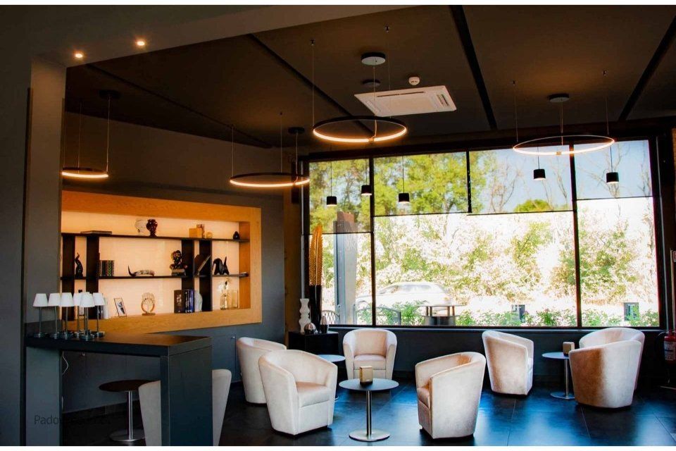 Area lounge con poltrone bianche, luci circolari, bar e ampie finestre con vista.