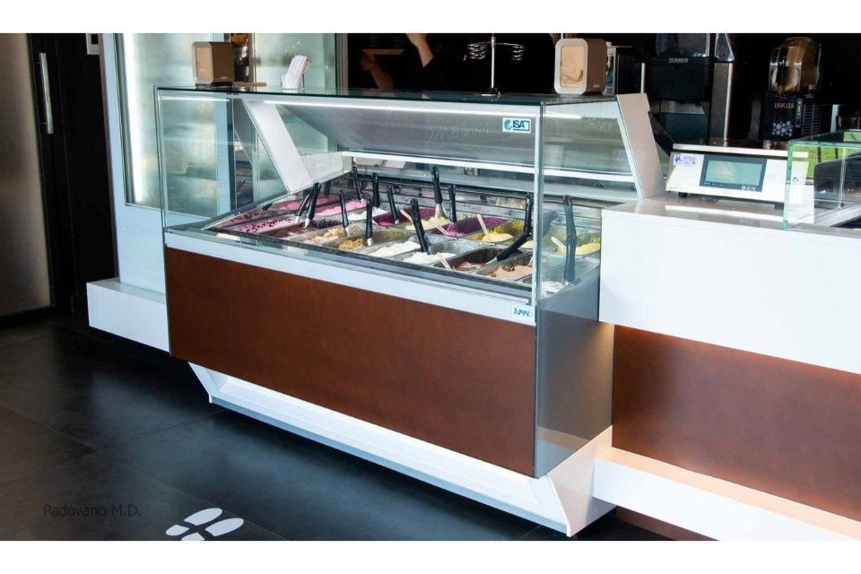 Vetrina per gelati con vari gusti colorati, facciata marrone e bianca, con pavimento nero.