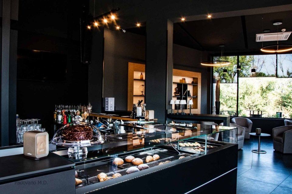 Interno di un bar buio con pasticcini, bevande e una grande finestra che si affaccia sul verde.