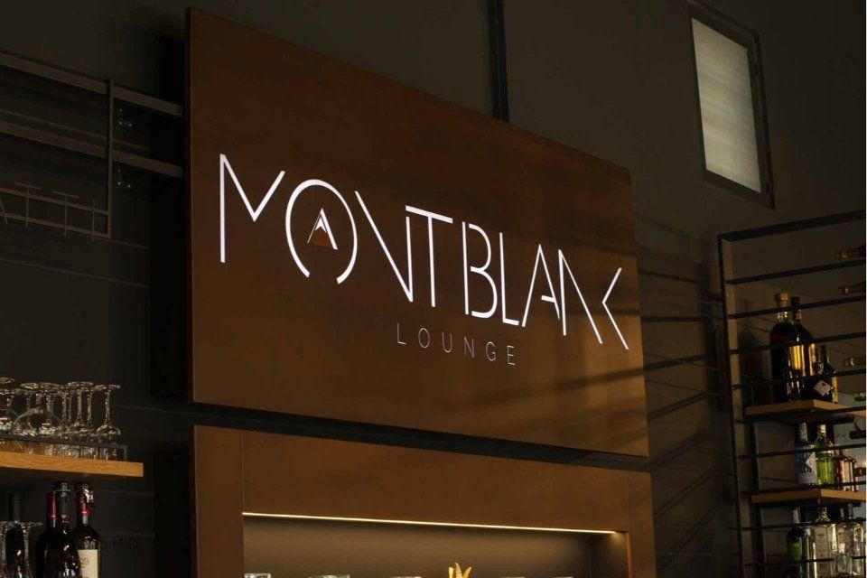 Insegna Montblanc Lounge, lettere bianche illuminate su parete marrone. Bar con bicchieri e scaffali a vista.