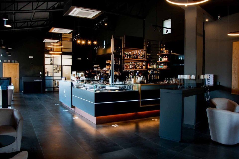 Interno del bar scuro con pareti nere, bancone retroilluminato e aree salotto.