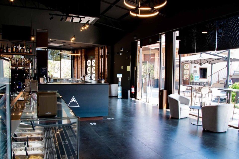 Vista interna di un bar moderno, con pareti e pavimenti scuri. Le porte a vetri si aprono su un patio esterno con tavoli e sedie.