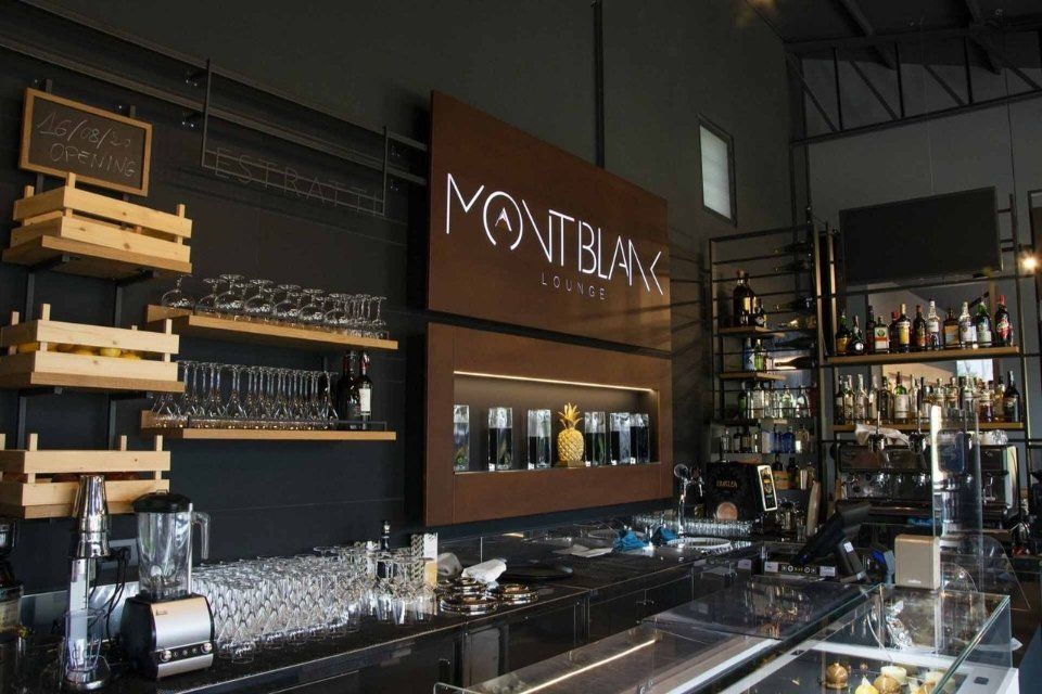 Vista interna del Montblanc Lounge bar. Con bancone, scaffali con bicchieri e bottiglie di liquore e logo su una parete marrone.