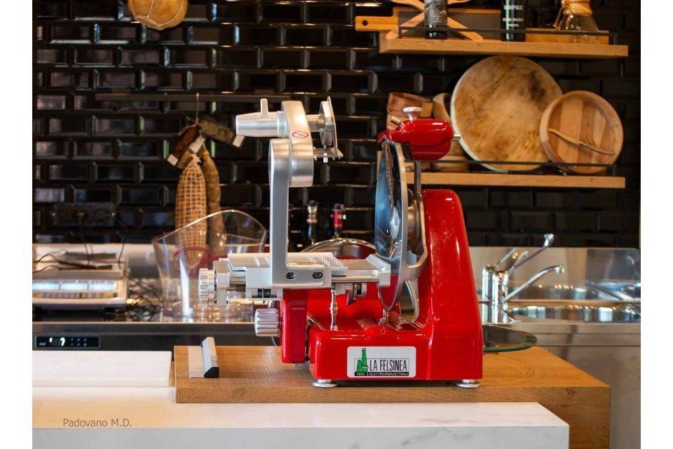 Affettatrice per carne rossa sul bancone di una cucina, con una parete piastrellata di nero e scaffali sullo sfondo.