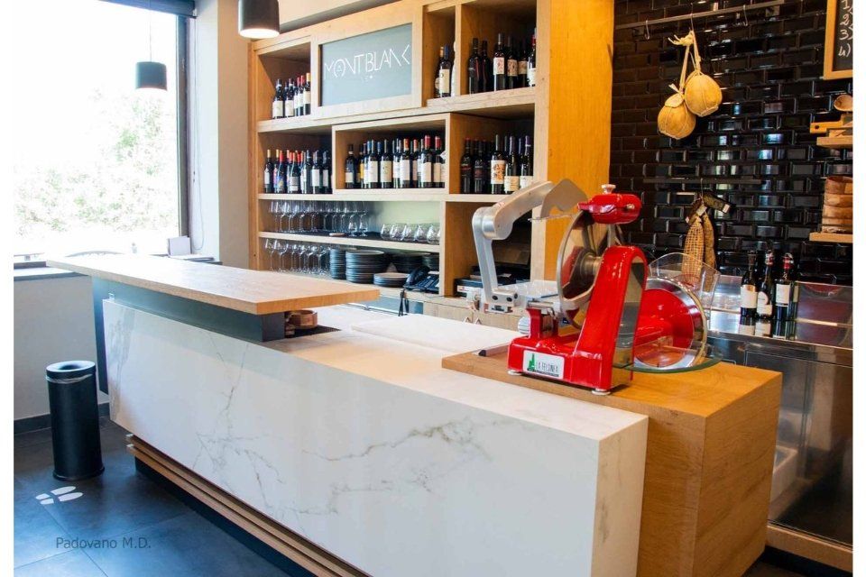 Interno di un bar con scaffali in legno pieni di bottiglie di vino, bancone bianco, affettatrice per carne rossa.