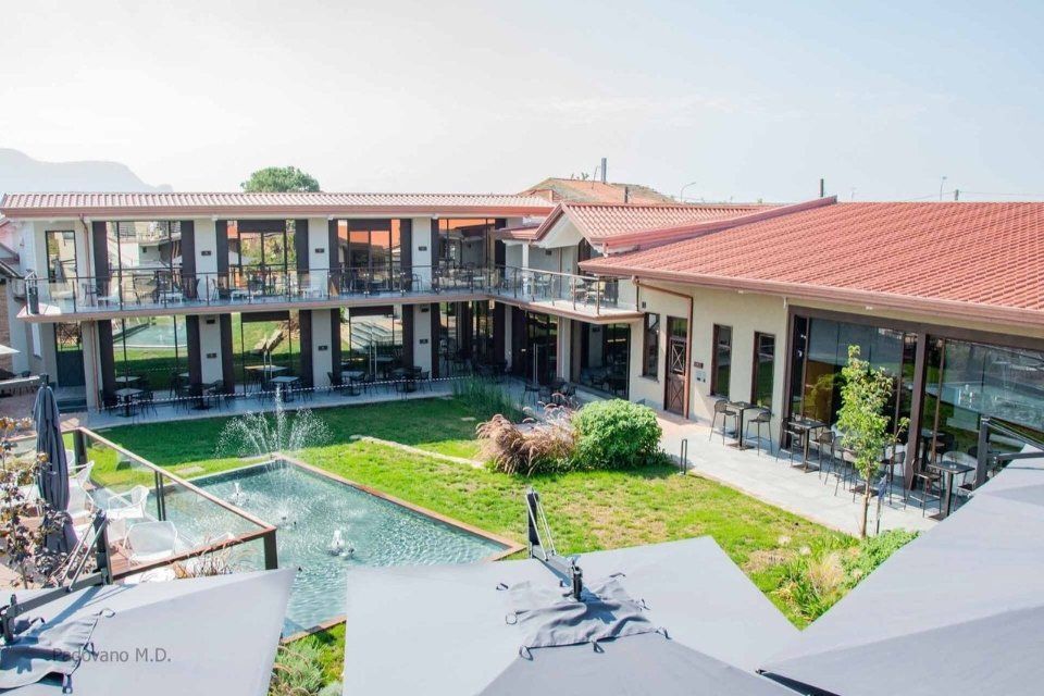 Hotel con cortile interno, edificio a due piani con tetto rosso. Fontana e prato verde al centro. Tavoli sui balconi.