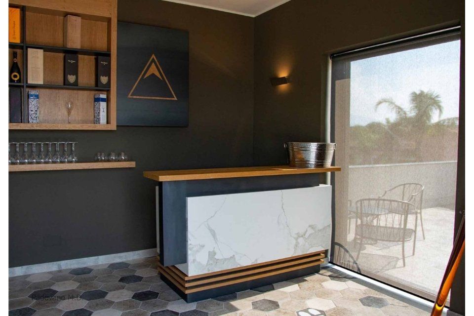 Bar con bancone in legno, facciata in marmo e mensola con bicchieri, accanto a posti a sedere all'aperto.