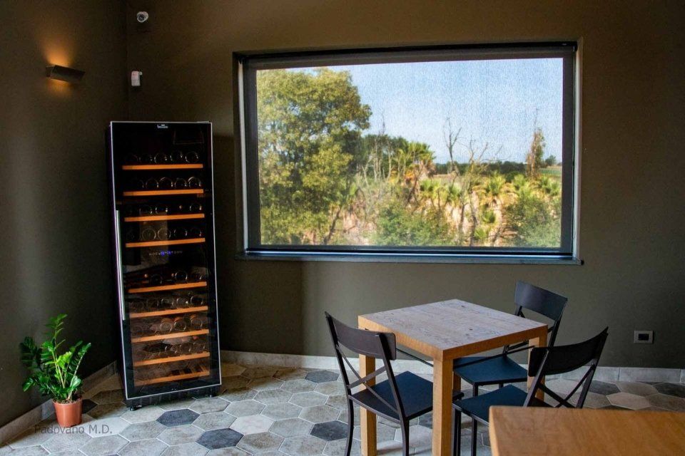 Mobiletto portabottiglie accanto a un tavolo quadrato con sedie nere, vicino a una finestra con vista sugli alberi.