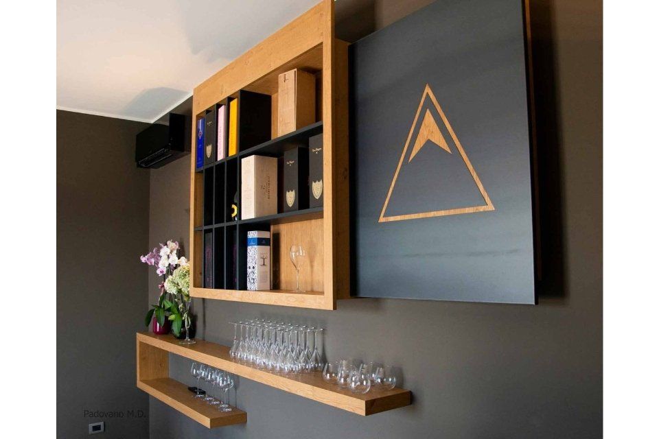 Scaffale in legno a parete con bicchieri da vino e scatole, accanto a una porta nera con il simbolo di un triangolo dorato.