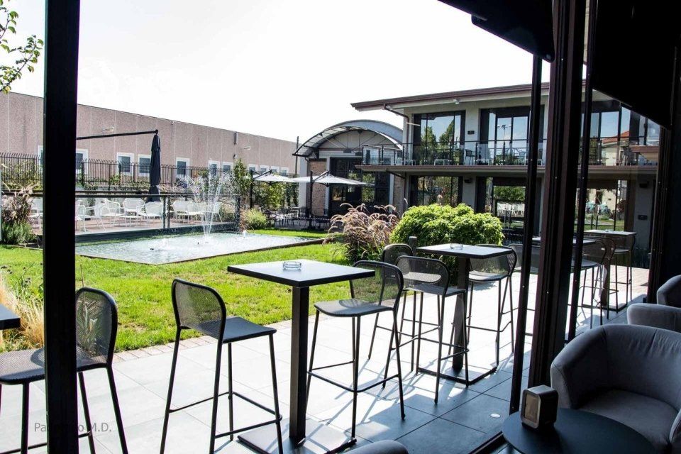 Vista del bar all'aperto con tavoli quadrati e sgabelli alti, con vista sul prato e sulla fontana.