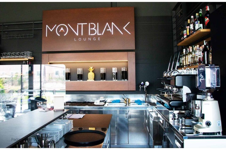 Interno della Montblanc Lounge. Area bar con macchina per caffè espresso, scaffali con bottiglie e insegna illuminata.