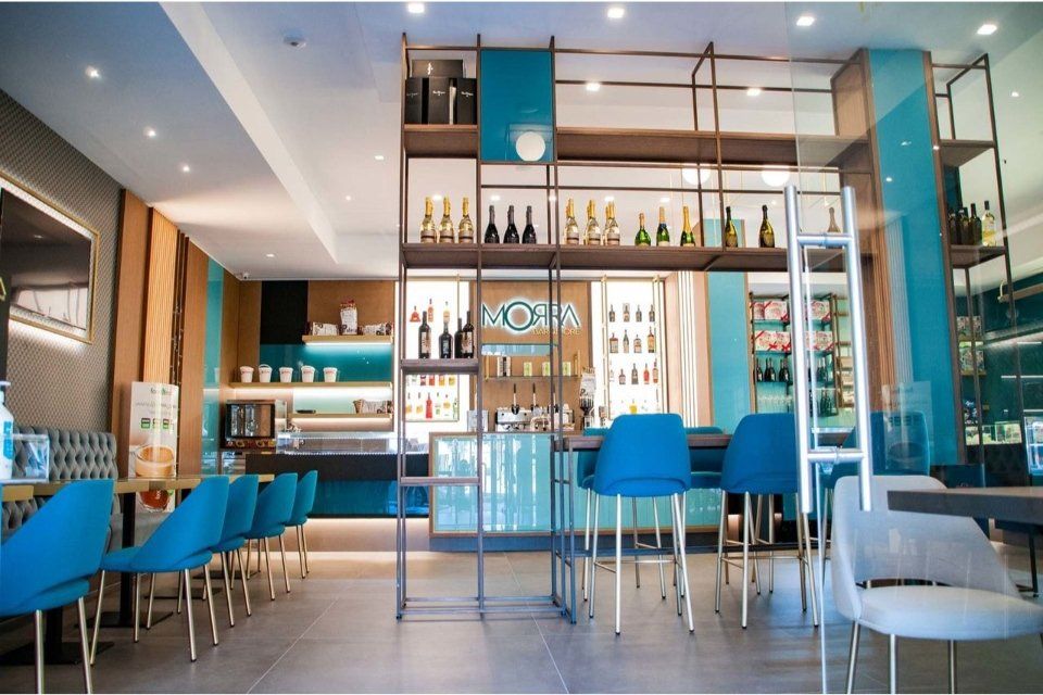 Interno di un moderno bar con dettagli turchesi, bancone, sgabelli e scaffali pieni di bottiglie.