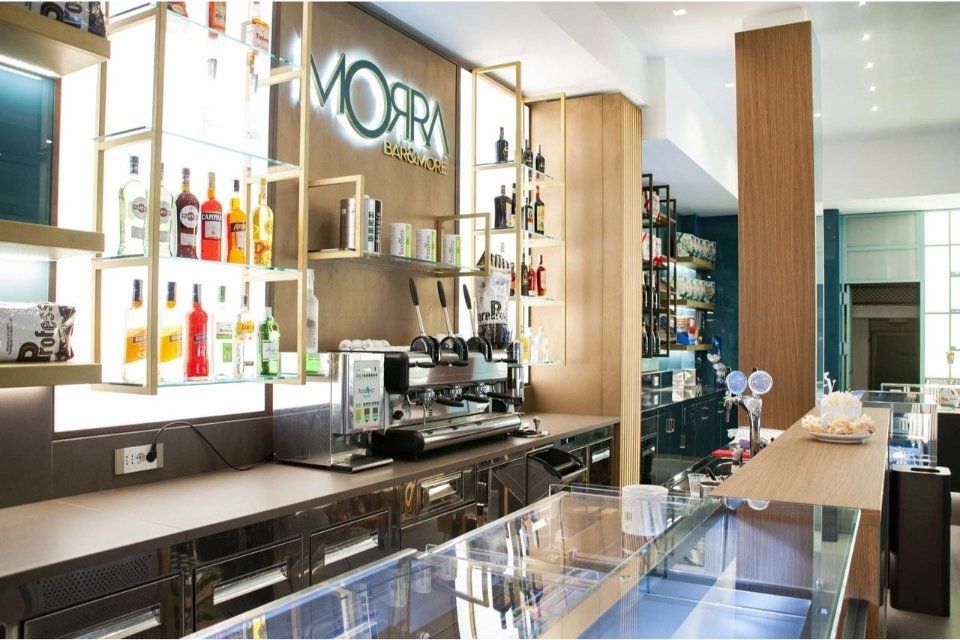 Interno del bar con scaffali illuminati che espongono bottiglie di liquore, macchina del caffè e bancone per il servizio. Insegna