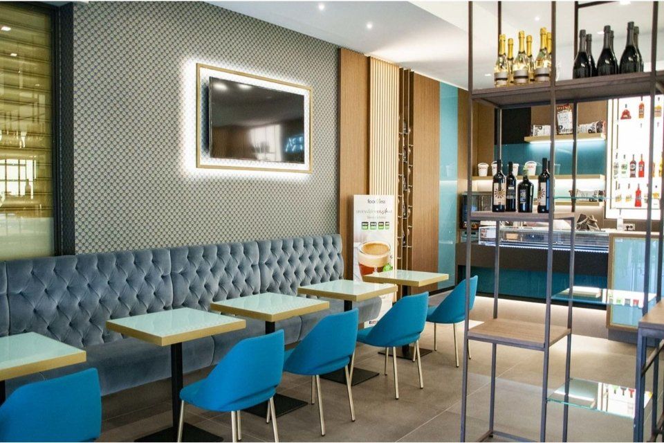 Interno del bar con tavoli e sedute blu, una televisione e scaffali con bottiglie in esposizione.