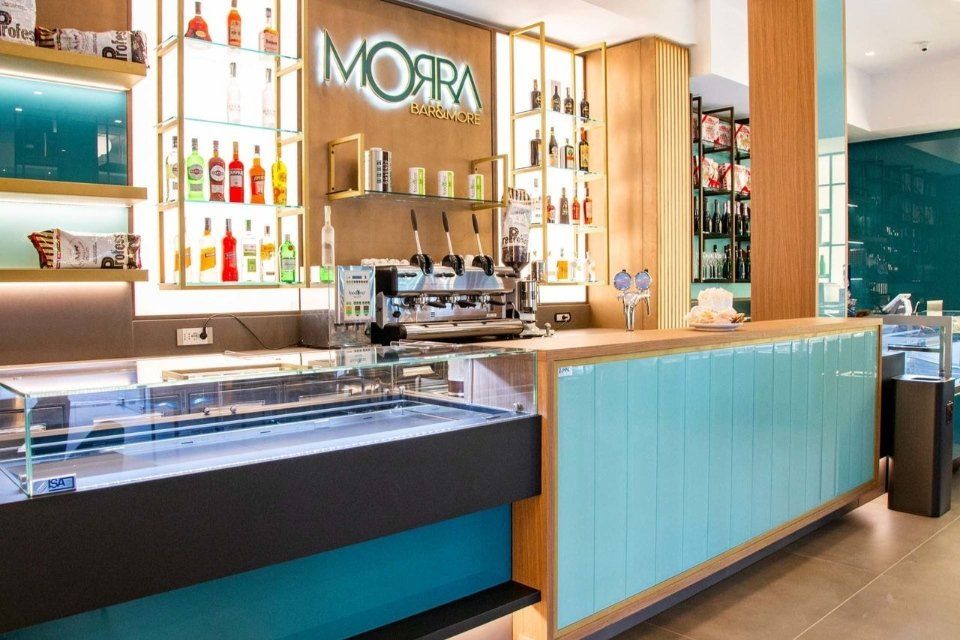 Interno del bar: toni azzurro e legno, bancone con bottiglie di liquore, macchina per l'espresso e insegna Moara.