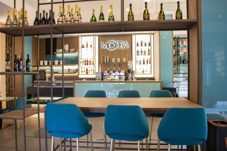 Interno di un ristorante con bar, posti a sedere e bottiglie sugli scaffali; tonalità verde acqua e legno.