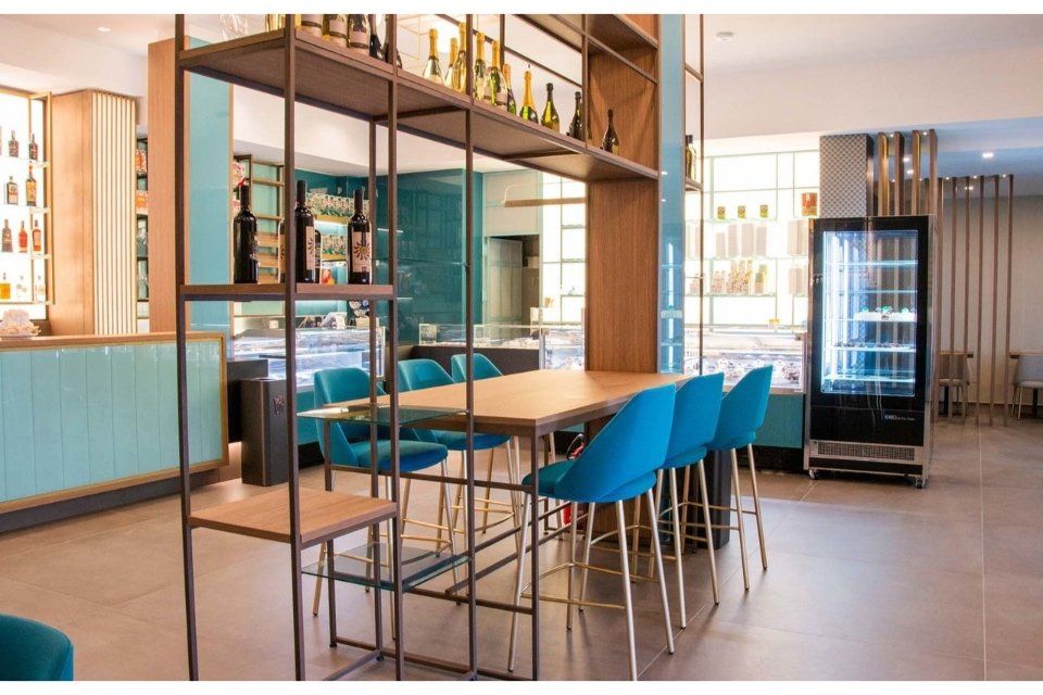 Interno di un bar moderno: sedie blu, tavolo in legno, scaffali con bottiglie e un frigorifero espositivo.