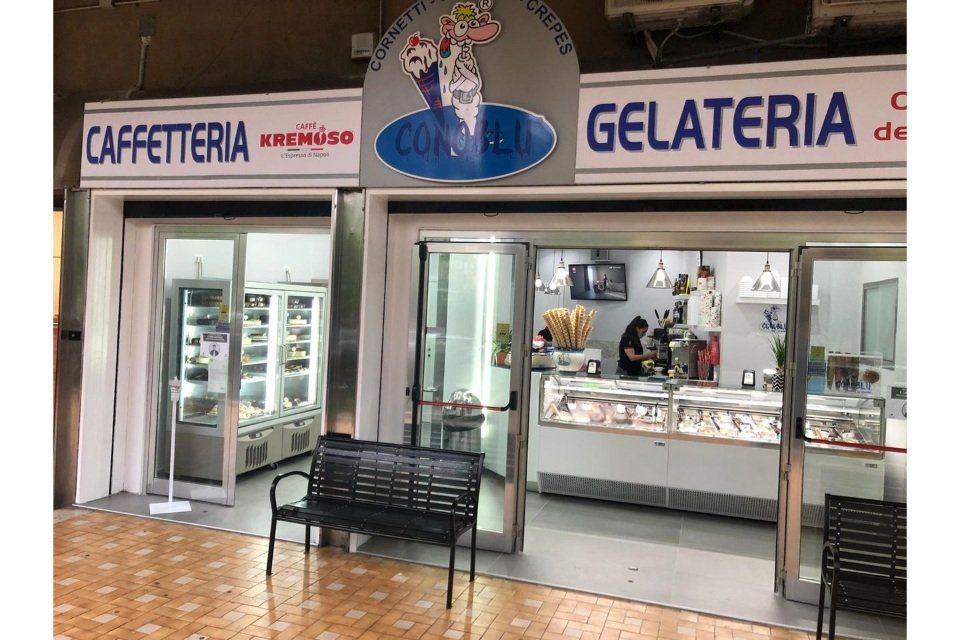 Facciata di un negozio di caffetteria e gelateria con porte in vetro, espositori e una panca nera.