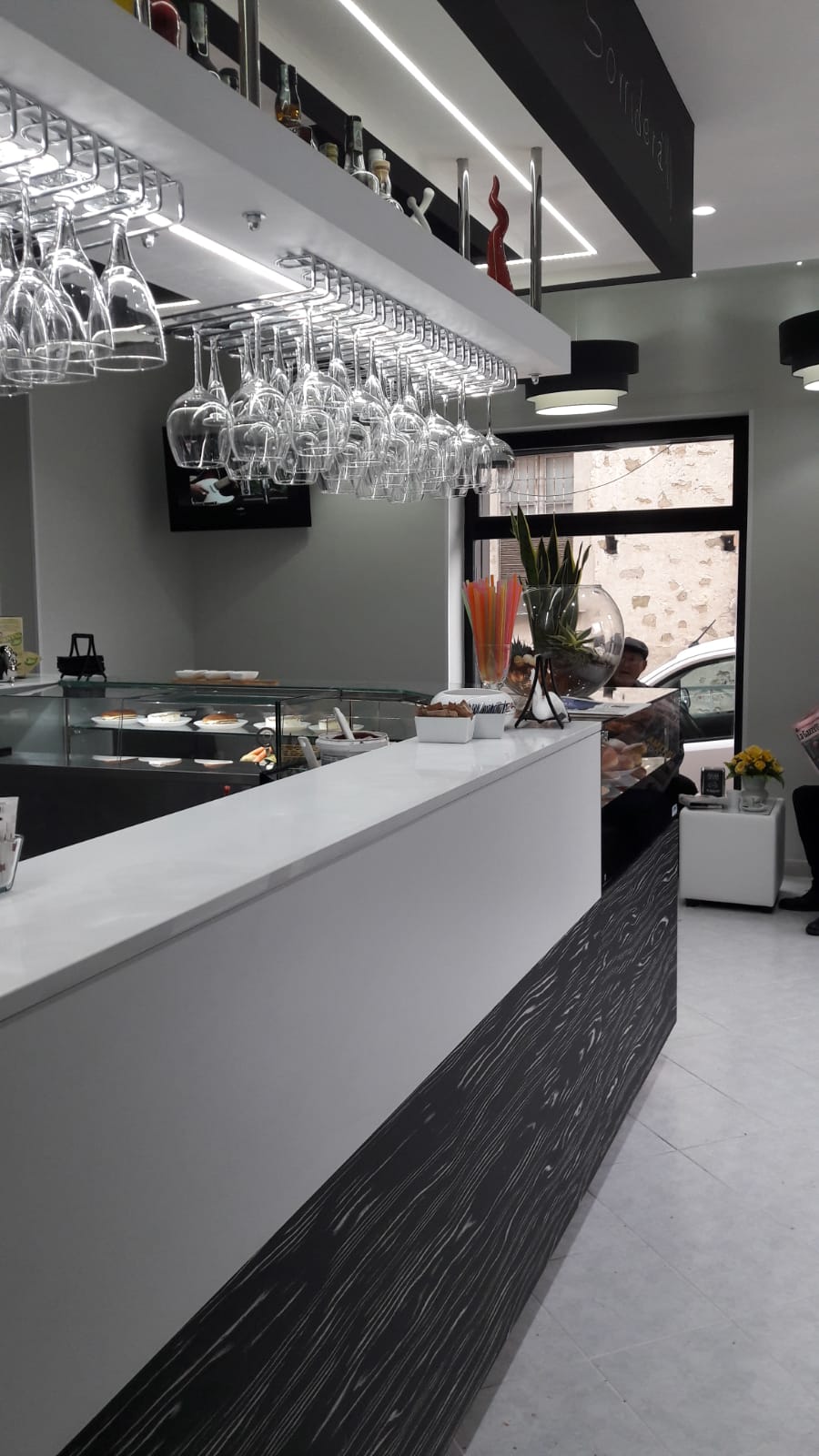 Interno di un bar con bancone bianco e bicchieri da vino appesi. È visibile un espositore di dolciumi.