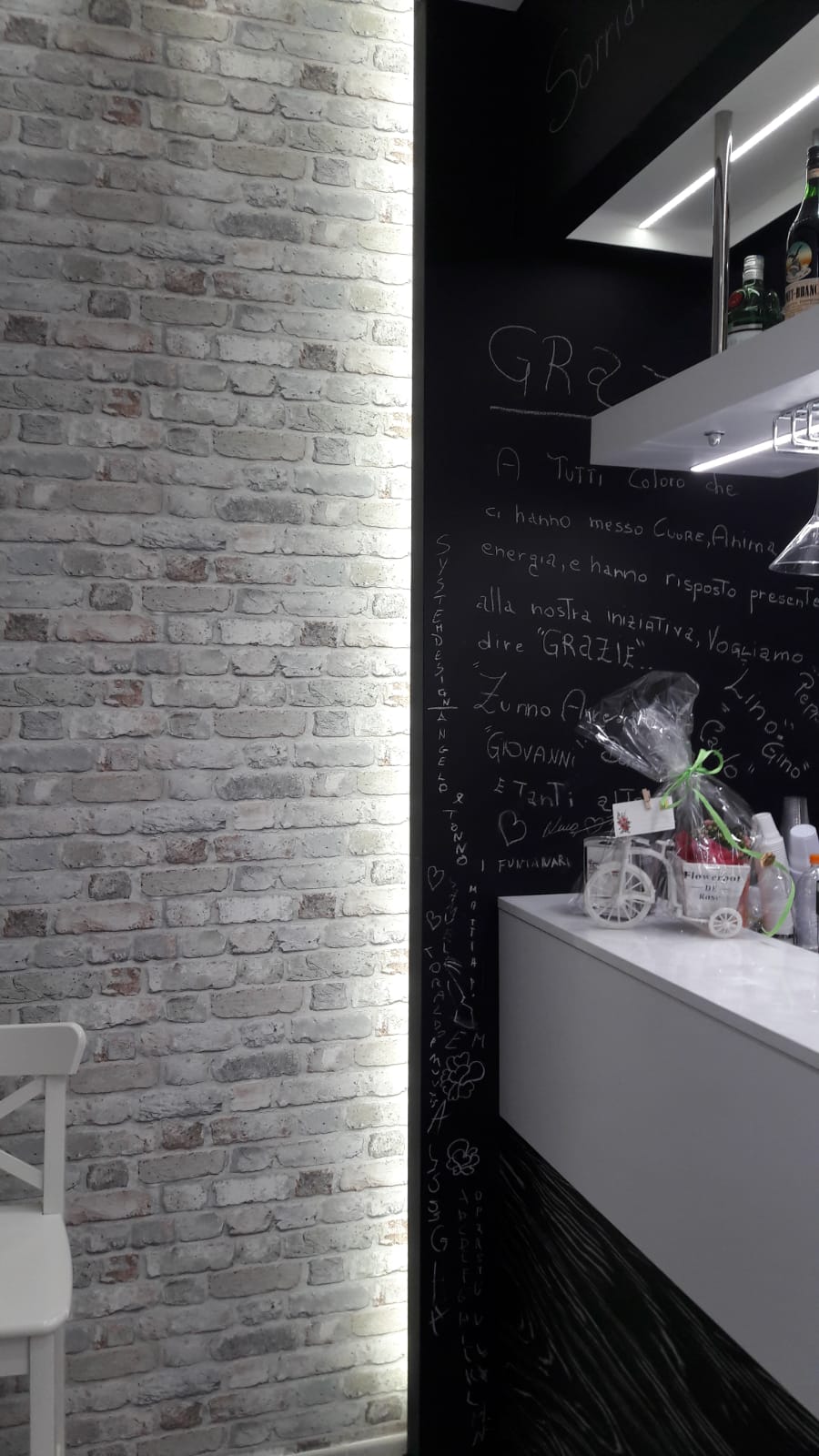 Parete in mattoni con striscia luminosa verticale, in contrasto con una parete scura in stile lavagna. Piano di lavoro e mensole bianchi.