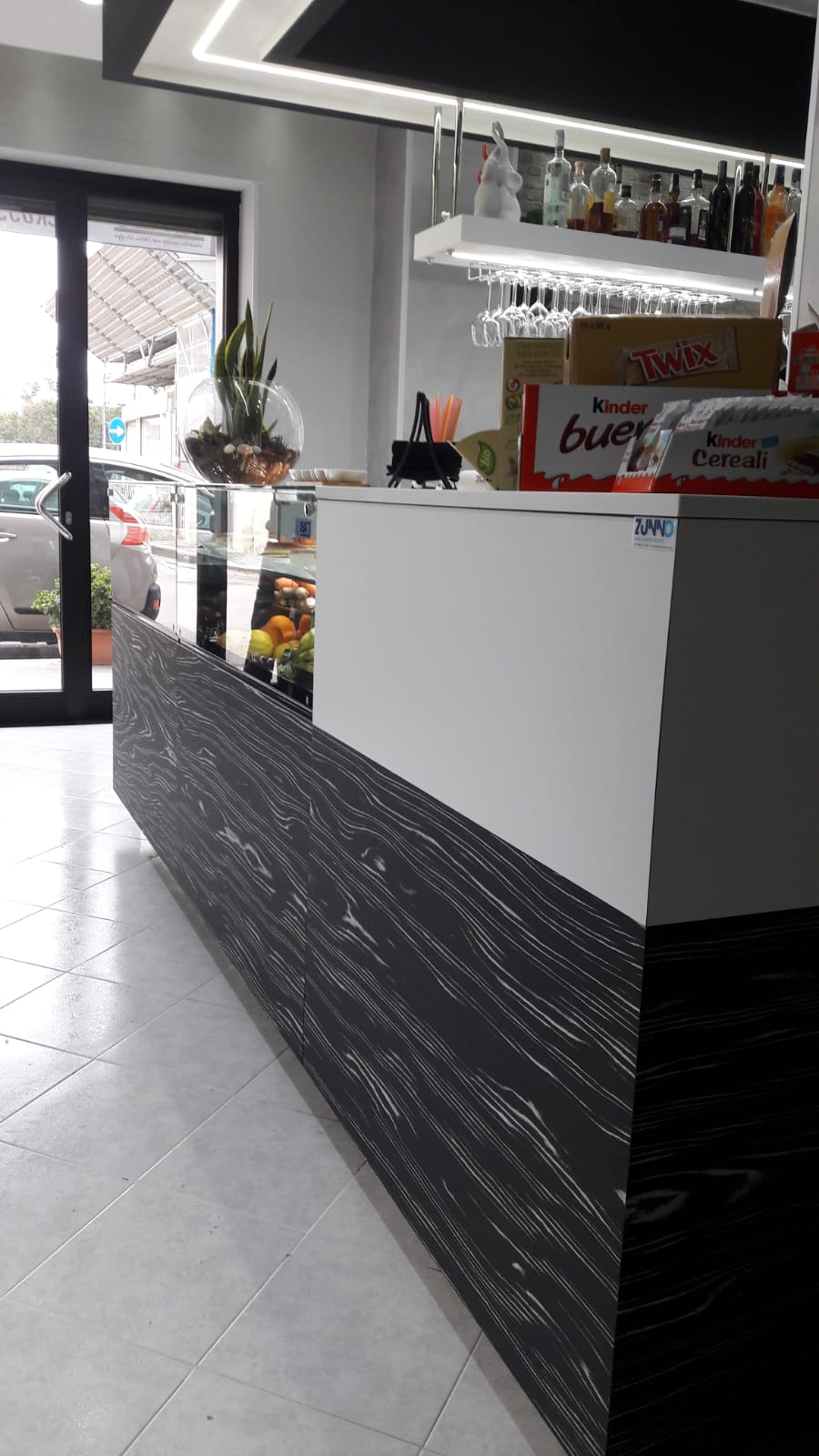 Interno di un bar con bancone in legno bianco e scuro. È visibile una porta a vetri, insieme a bevande e pasticcini.