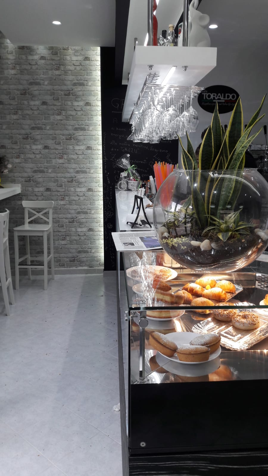 Interno di un bar con dolci esposti in una vetrina, un muro di mattoni bianchi e posti a sedere alti al bancone.