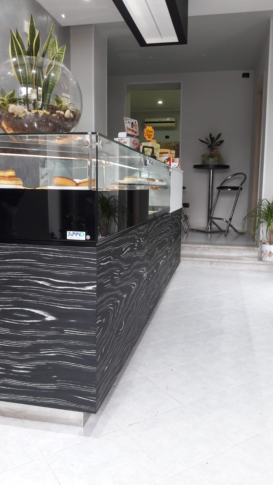 Bancone da panetteria con pasticcini. Facciata in marmo bianco e nero. Piante in una ciotola di vetro. Interni luminosi.
