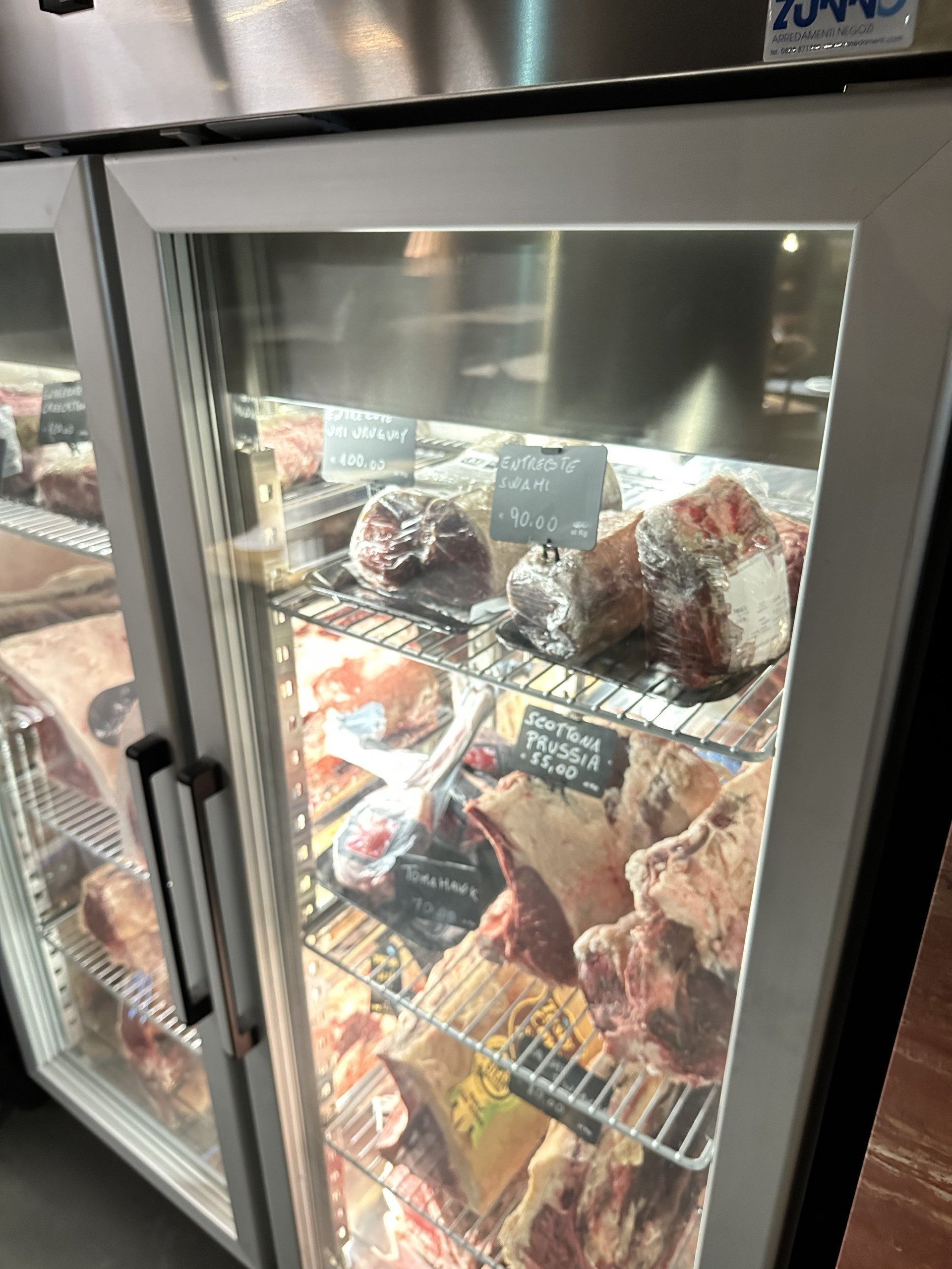 Vetrina con tagli di carne frollata a secco. Porte in vetro, ambiente refrigerato, etichette visibili.