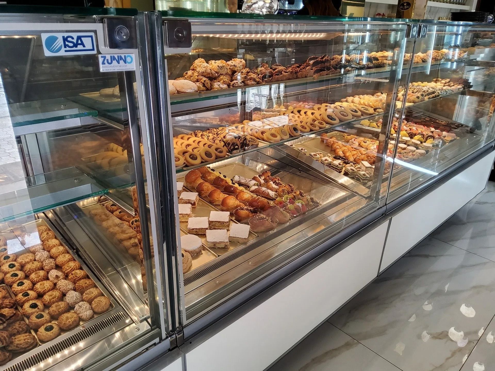 Vetrina da pasticceria piena di vari tipi di pasticcini e biscotti. Base bianca, ripiani in vetro e illuminazione intensa.