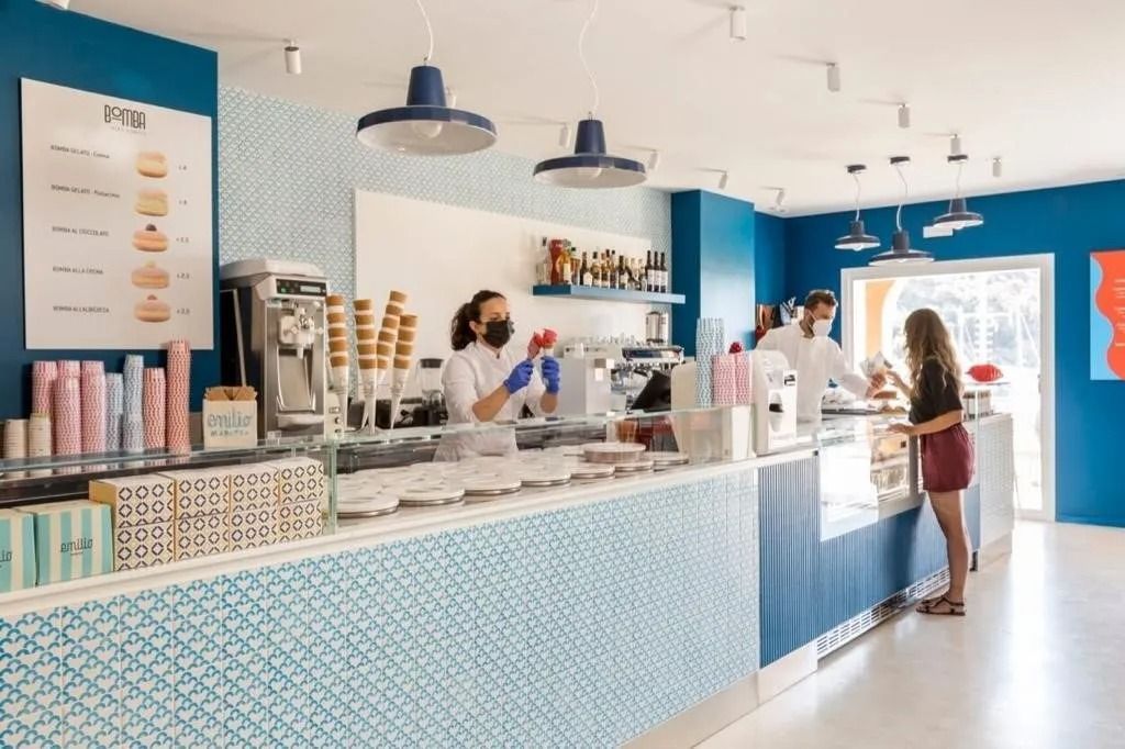 Interno di una gelateria con clienti e personale dietro al bancone. Decorazioni blu e bianche.