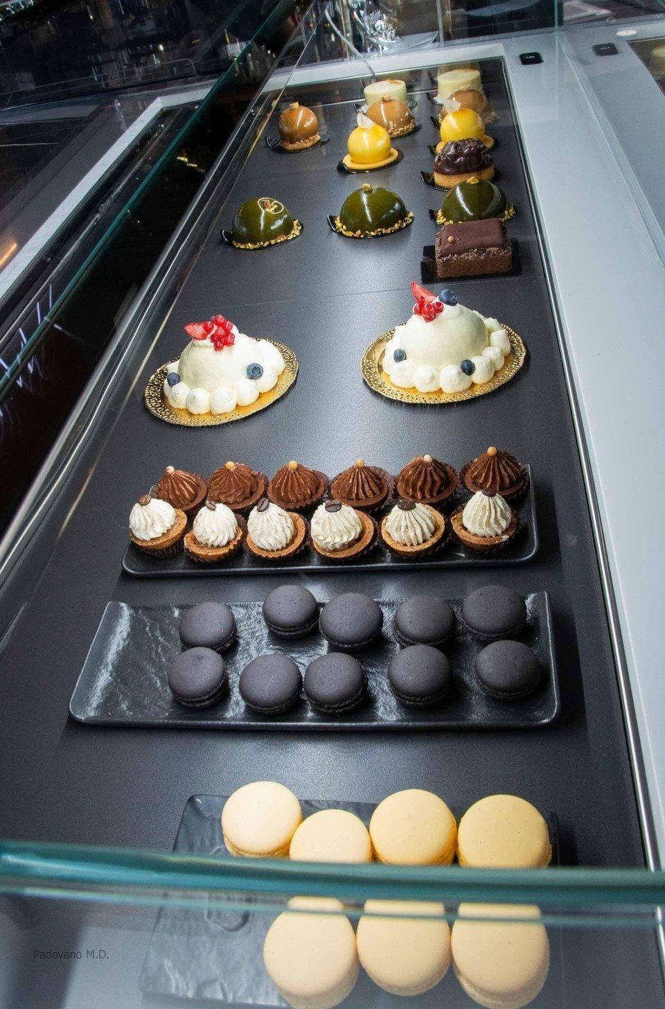 Vetrina con vari tipi di pasticceria: torte glassate, crostate e macaron nei toni del bianco, verde, marrone e giallo.