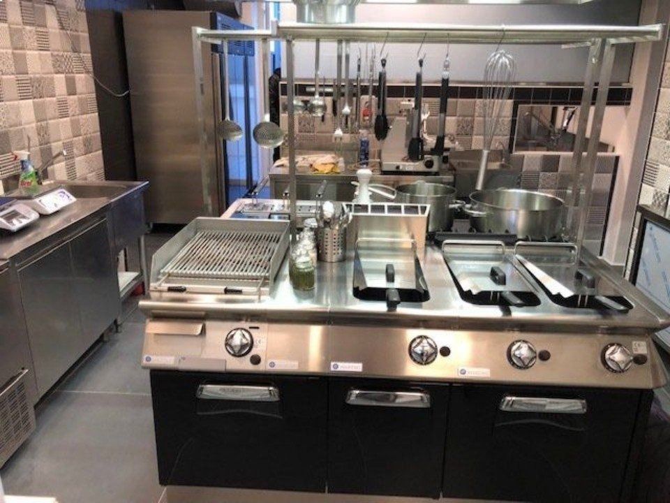 Cucina commerciale con elettrodomestici in acciaio inossidabile: grill, friggitrice, piano cottura e utensili appesi.