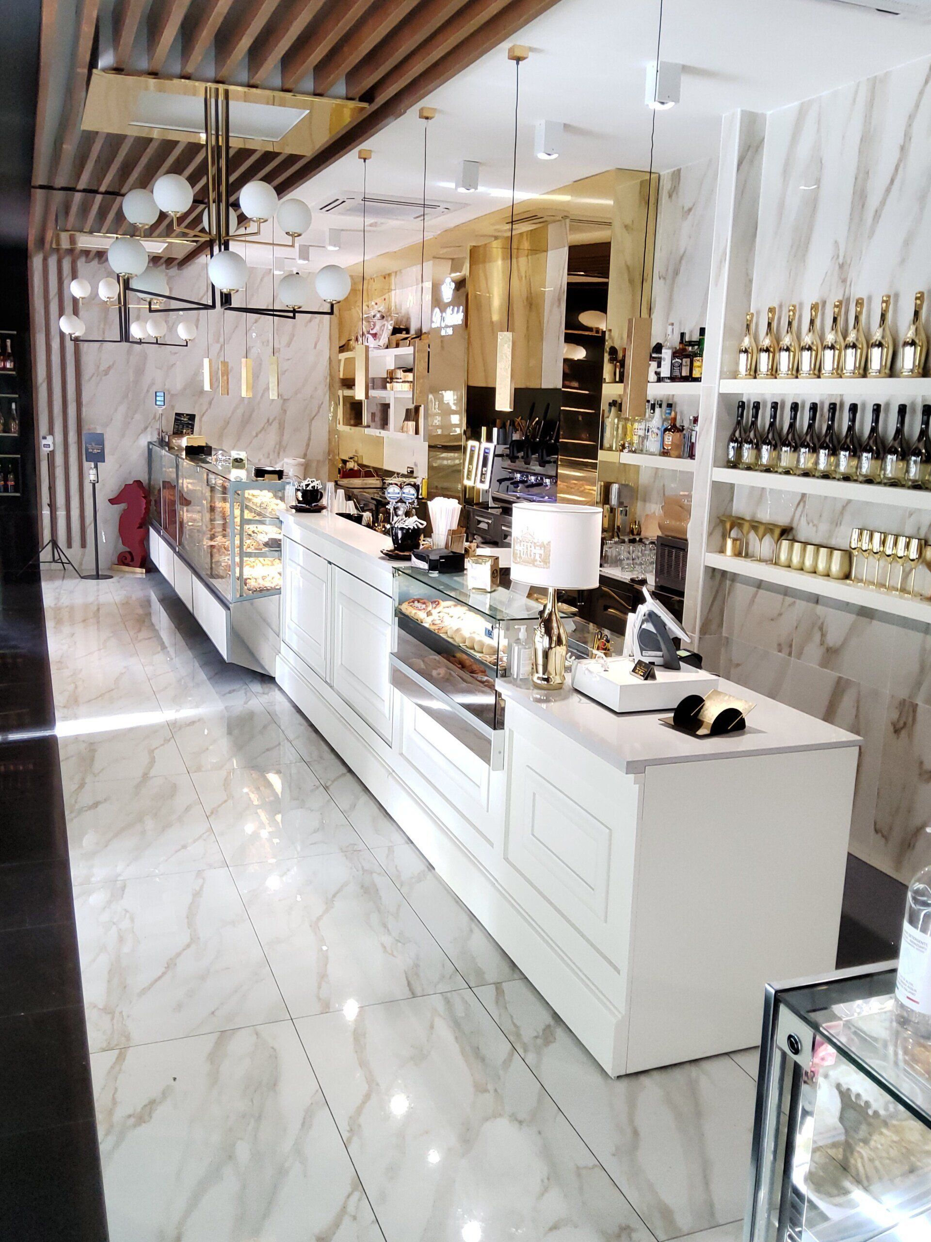 Una pasticceria ben illuminata, bianca e dorata, con vetrine e prodotti esposti sugli scaffali. Pavimento in marmo.