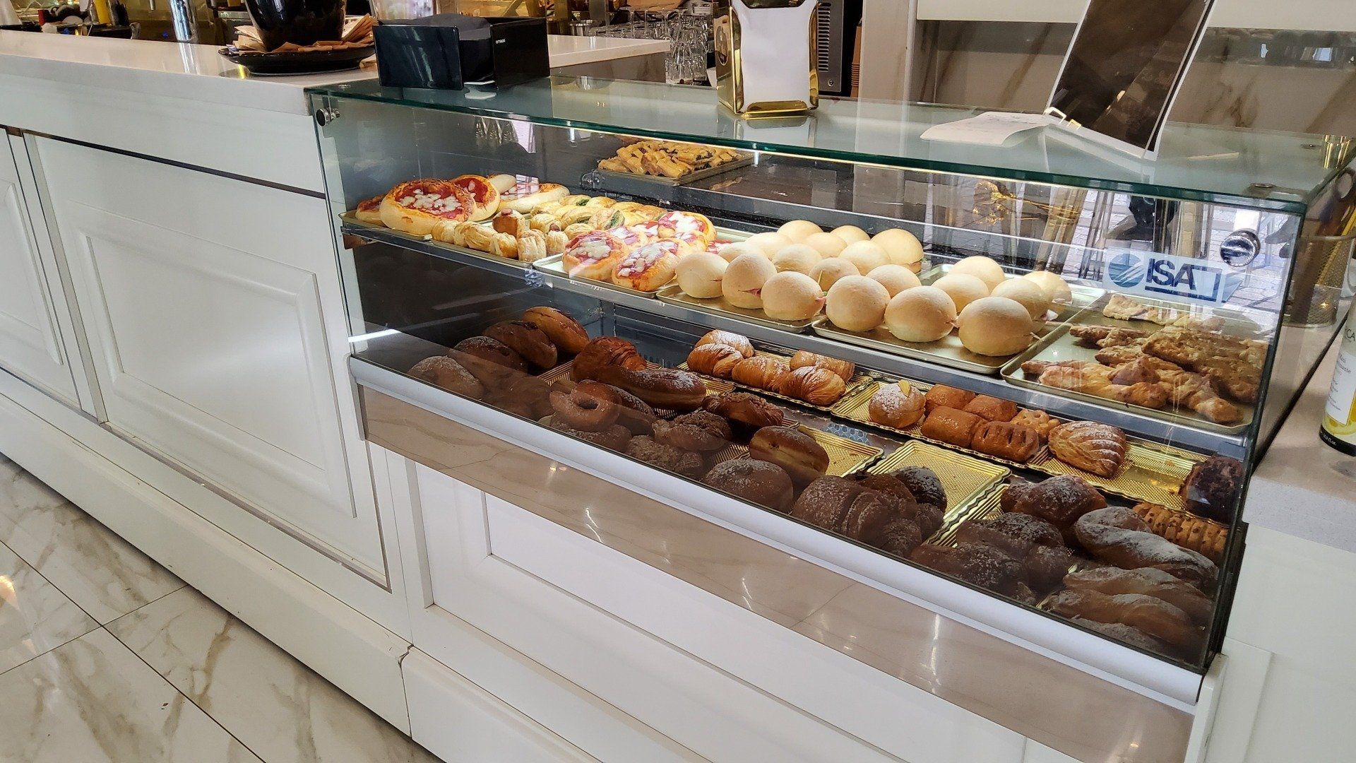 Vetrina da panetteria piena di dolci: croissant, panini, pizza e pane. Mobili bianchi e pavimenti in marmo.