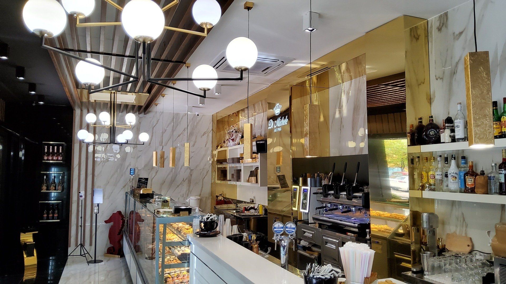 Interno di un bar arredato in bianco e oro, con bancone e lampade a globo sospese.