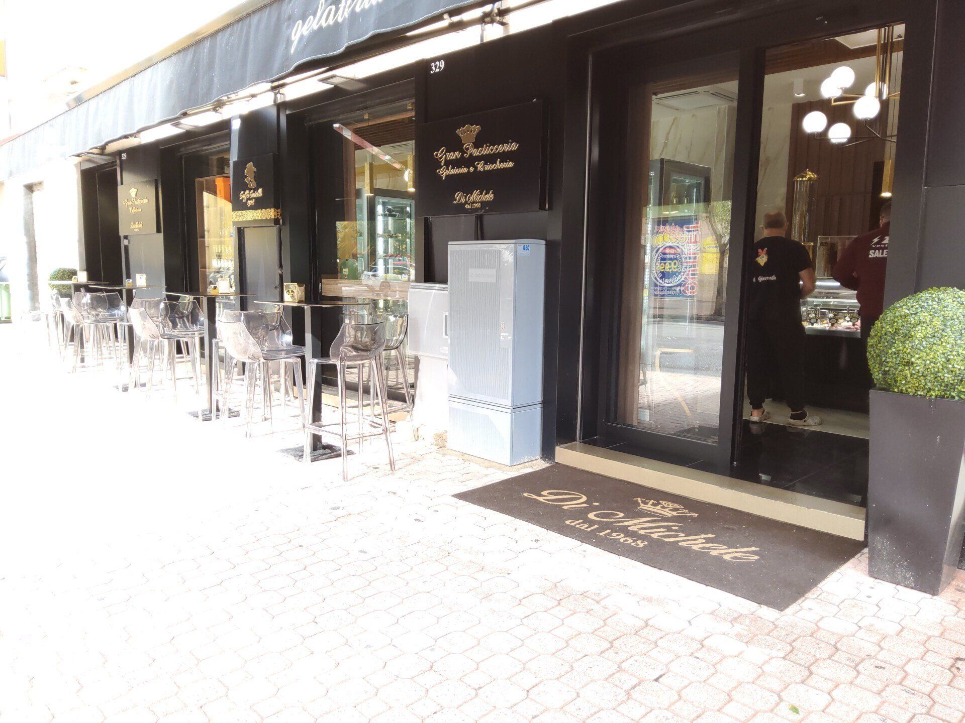 Esterno del bar con sedie trasparenti, tenda da sole e zerbino.