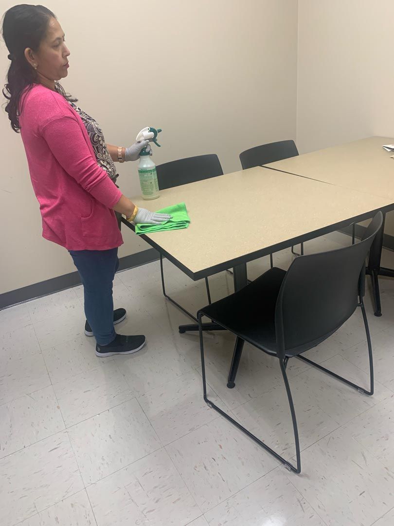 A woman in a pink shirt is cleaning a table with a spray bottle.