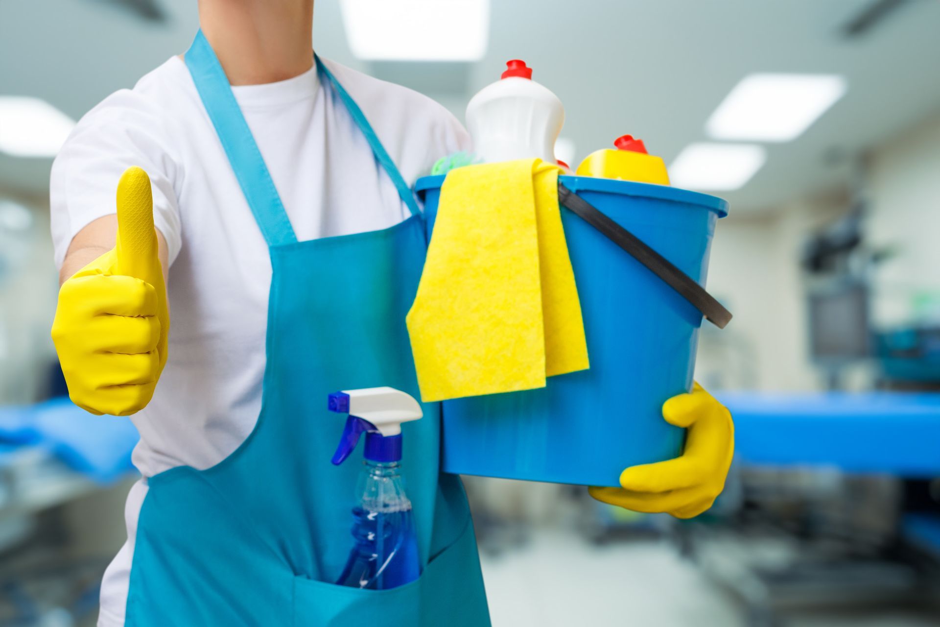 Person in apron holding cleaning supplies, giving thumbs-up in a commercial setting.