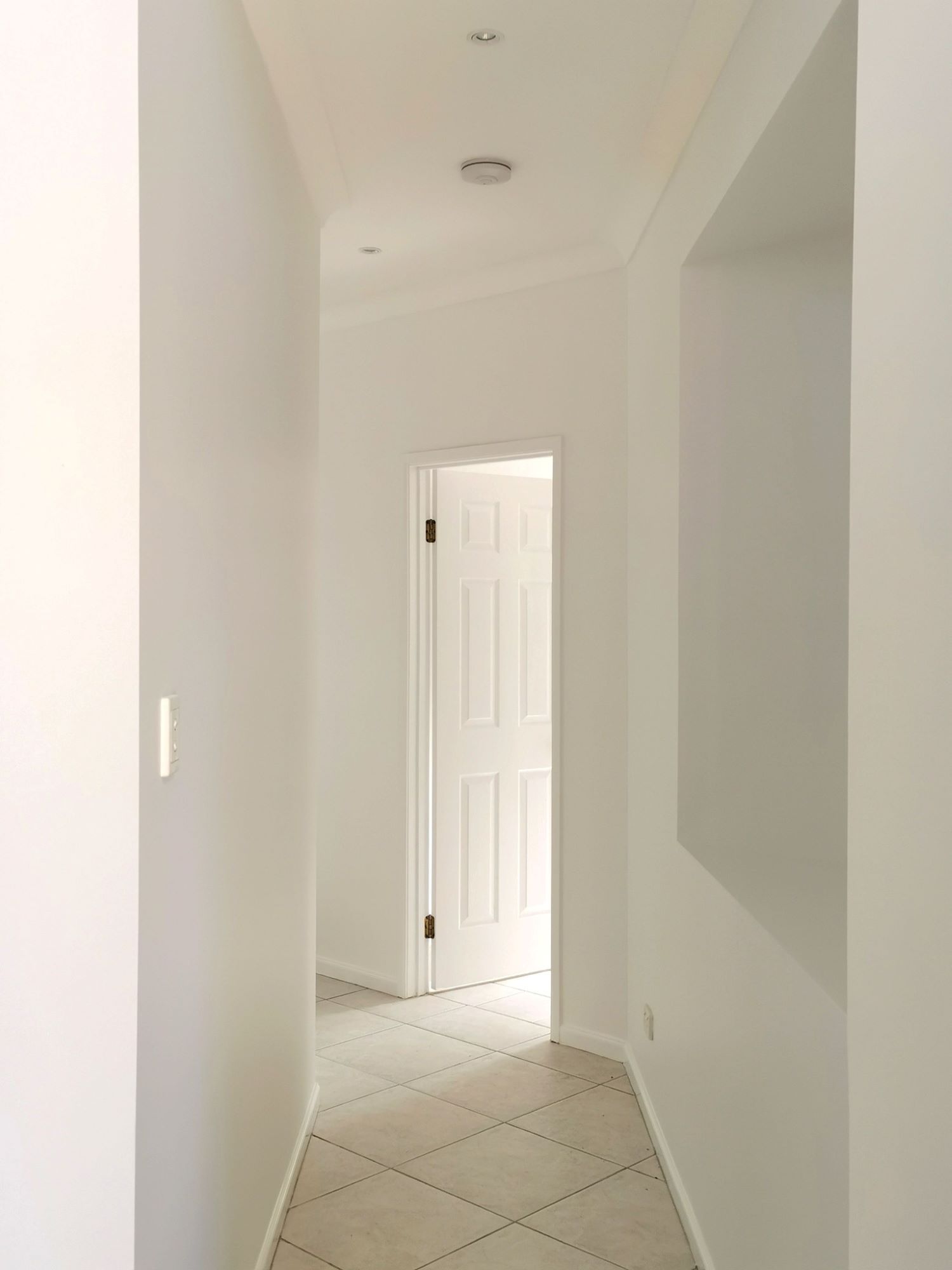 A Long Hallway With White Walls and Tile Floors Leading to a Door — Eva Professional Painting in Palm Beach, QLD
